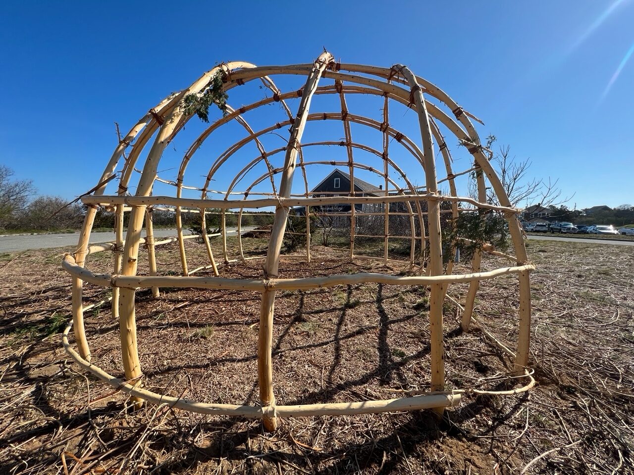 A wetu structure stands against a bright blue sky.