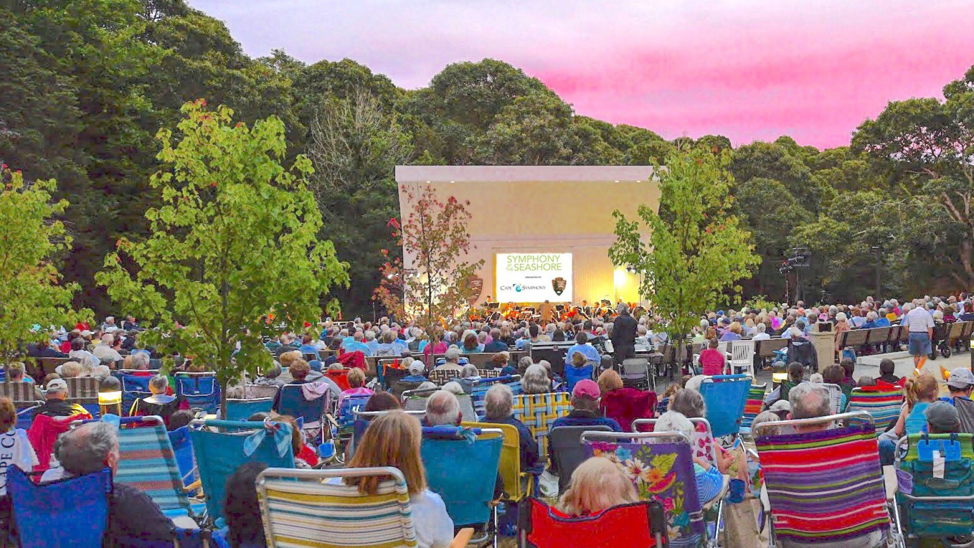 Large group of visitors sits in chairs facing a stage against a sunset sky.